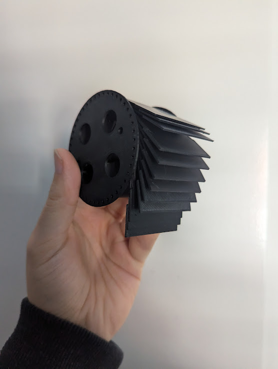 A split flap rotor with some flaps installed. All plastic parts are printed in black plastic. The object is being held in a hand.